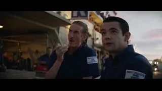 two men in blue polo shirts by Kia sign — Ford v Ferrari